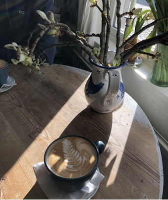 A cappuccino in a cup resting on a table in front of a sunny window.